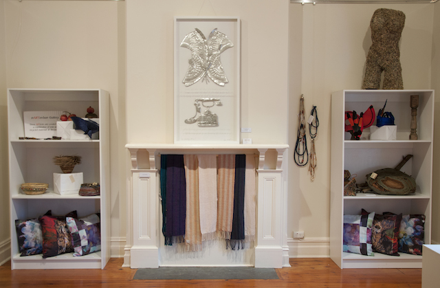 North wall of Annex, showing basketry, felt necklaces, weaving and cushions and above the mantle Karen Genoff’s This Suffocating Silence in embossed aluminium, glass and text, $3000.